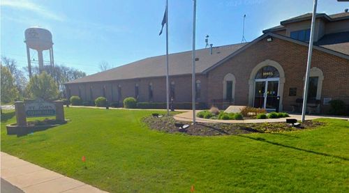 Front of the Town Hall building for the town of St. John Indiana with the sign in the grass with text "St. John Town Hall" and the St. John Indiana town water tower in the background
