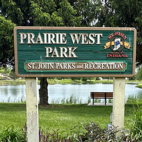 Park sign for Prairie West Park in St. John Indiana. It is a wood sign with green background with the text of "PRAIRIE WEST PARK St. John Parks and Recreation" in white font and the logo for the town of St. John is also on the sign.
