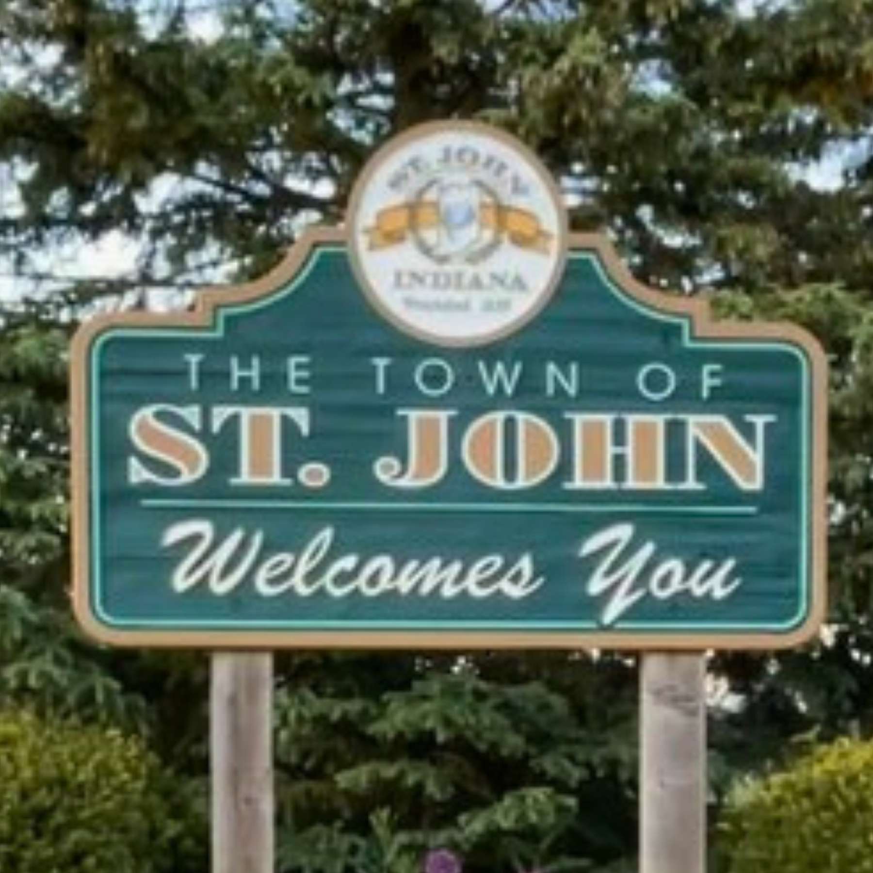 Town of St. John Indiana roadside entrance sign with text of "The Town of St. John Welcomes You" with an evergreen tree in the background